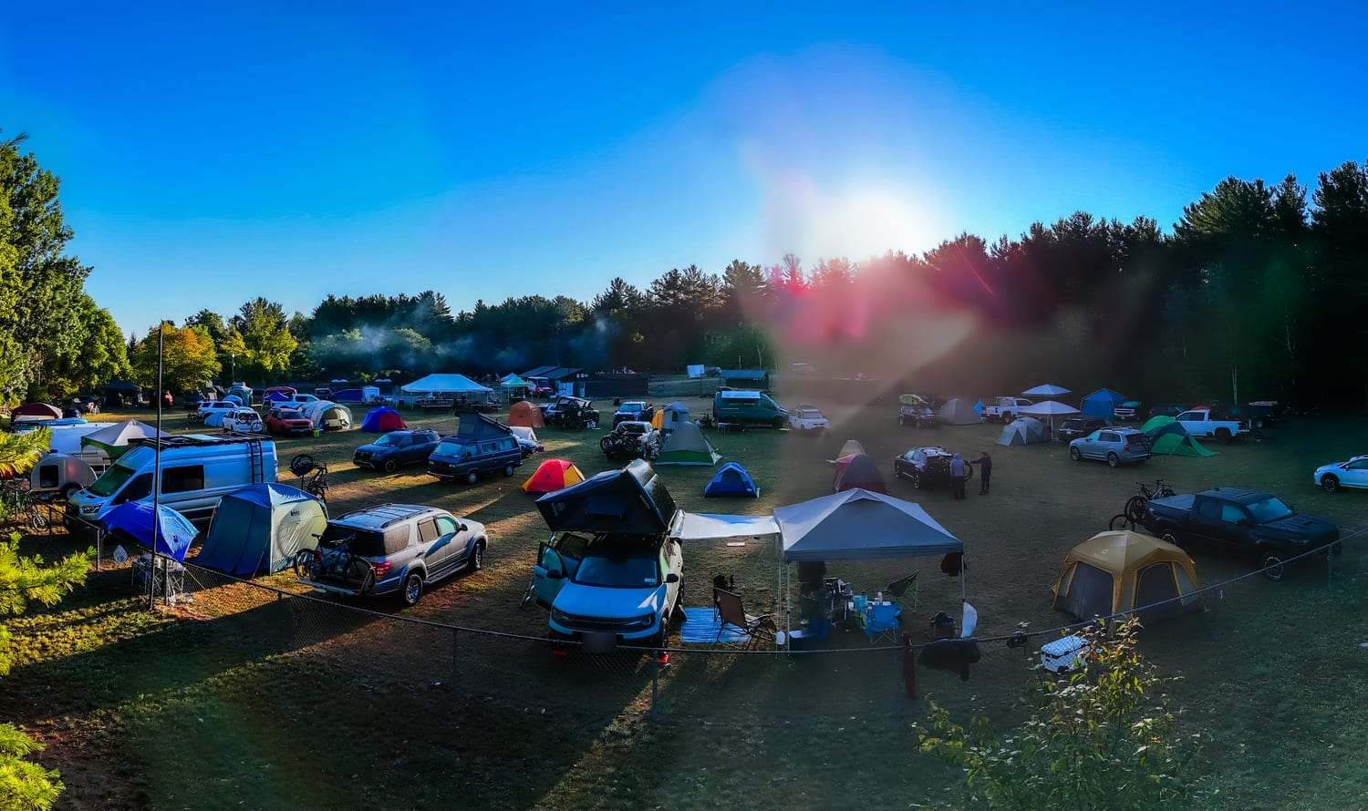 Tents, roof tents, vans and trucks spread across a grass field at the edge of a pine forest, with low sun behind the trees