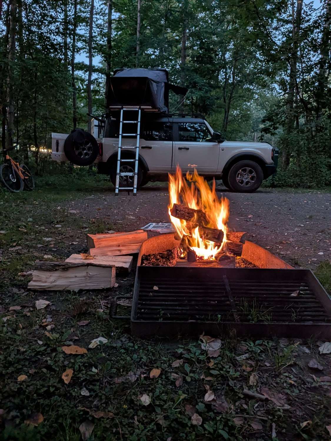 A campfire burning in a fire ring at dusk, with a Bronco under an open roof tent and a mountain bike behind it