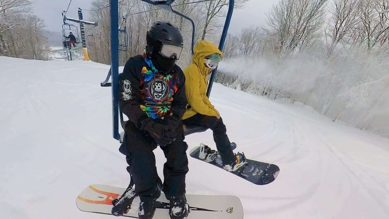 A snowboarder on a chairlift beside a rider in a yellow hood