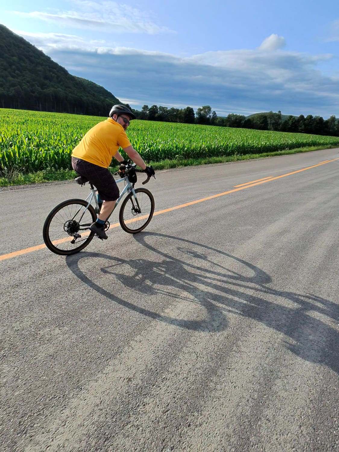 A rider in a yellow jersey on a gravel bike, on a country road beside a green corn field