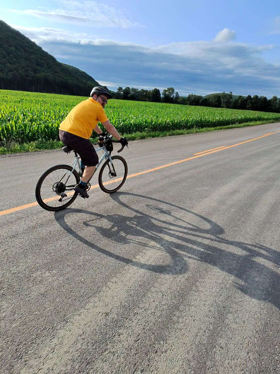 A rider in a yellow jersey on a road beside a green corn field